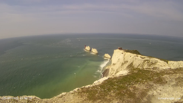 A panoramic view of dramatic white chalk cliffs rising sharply from the sea, with a grassy top and a small building on the edge. In the distance, tall white sea stacks stand in the water, with one featuring a red lighthouse. The ocean is a mixture of green and blue, with gentle waves and a few sailboats far away on the horizon. The sky is partly cloudy with soft, wispy clouds. The scene appears to be taken during daytime with clear, bright weather.
