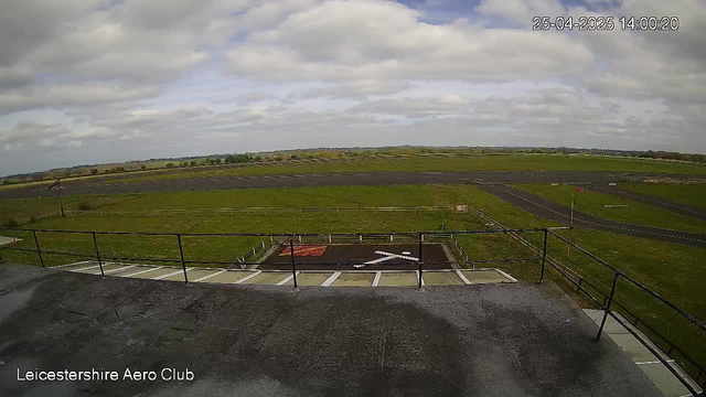 A wide, open landscape view from a webcam at Leicester Shire Aero Club. The foreground shows a concrete platform with a black railing, and a small helipad marked with a white arrow. Beyond the platform, there are grassy fields stretching toward the horizon, with a few scattered trees and hedgerows. The sky is partly cloudy with patches of blue. The timestamp in the top right corner reads "25-04-2025 14:00:20".