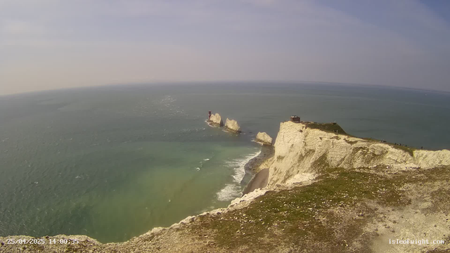 Aerial view of white chalk cliffs stretching along the coastline with a grassy top, overlooking the ocean. In the distance, a small lighthouse stands on a rock formation surrounded by water. The sky is partly cloudy with a gentle horizon line where the sea meets the sky. The water near the shore is a light green, gradually blending into deeper blue further out. There is a timestamp in the bottom left corner indicating the date and time.