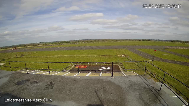 An empty helipad with a large white "H" symbol and a red square, surrounded by a black railing, located at Leicestershire Aero Club. The scene overlooks an open grassy area with a runway in the background under a partly cloudy sky. The timestamp shows "25-04-2025 15:00:11" in the top right corner.