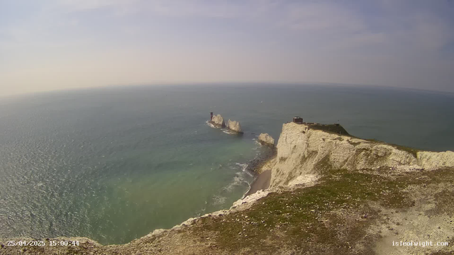 A view of a coastline with white chalk cliffs extending into the ocean. There are two small islets with a lighthouse on one of them. The sky is mostly clear with a few clouds, and the water has gentle waves. The photo is timestamped "25/04/2025 15:00:44" and has a website watermark "isleofdight.com" in the bottom right corner.