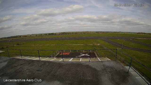 An expansive view of an airfield under a partly cloudy sky, with runways and green grassy areas stretching into the distance. In the foreground, there is a small helipad marked with an "H" and surrounded by safety barriers, with a few white lines indicating parking or loading zones. The bottom left corner has the text "Leicestershire Aero Club." The timestamp in the top right corner reads "25-04-2025 16:00:39."