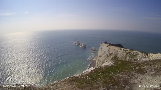 A panoramic view of white chalk cliffs along the coast with a small building on top, overlooking the ocean. The sea appears calm with gentle waves, and the sky is clear with a few thin clouds. In the distance are rock formations rising from the water. The image is taken in bright daylight, showcasing the scenic landscape.