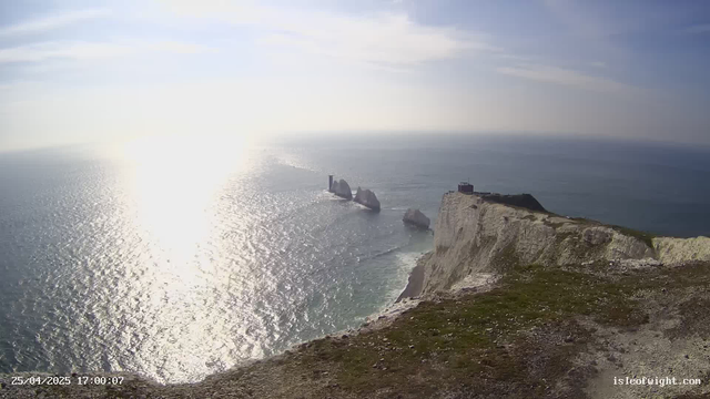 A panoramic view of a coastal landscape featuring tall white chalk cliffs with patches of green grass on top, extending into the ocean. Several large sea stacks rise from the water near the base of the cliffs, and a lighthouse is visible on one of the stacks. The sea is shimmering under sunlight, and a small building is situated on the edge of the cliff. The sky is partly cloudy with soft light, creating a serene atmosphere. The timestamp in the bottom left corner reads "25/04/2025 17:00:07," and the bottom right corner has the website "isleofwight.com."