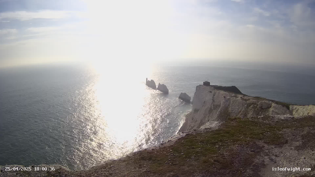 A view of white chalk cliffs along the coastline with a grassy top, extending into the distance. The sun is low in the sky, creating a bright reflection on the ocean below. There are a few small structures on the edge of the cliffs. The date and time stamp in the bottom left corner reads "25/04/2025 18:00:36," and the bottom right corner displays "isleofwight.com."