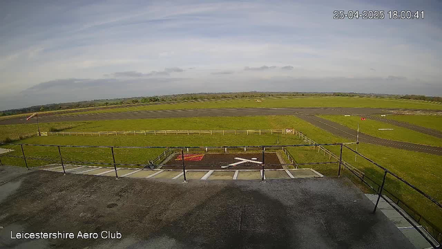 An aerial view of an open green field with a runway extending into the distance, under a partly cloudy sky. In the foreground, there is a small parking area with marked parking spaces and a metal railing, labeled "Leicestershire Aero Club" in the bottom left corner. A windsock and runway lights are visible, indicating this is an airport or airfield. The timestamp shows the date and time as April 25, 2025, at 18:00:41.