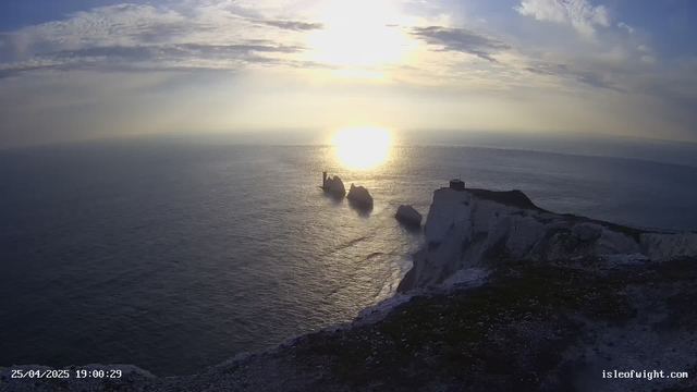 A view of white chalk cliffs overlooking the ocean at sunset, with the sun low in the sky casting a golden light. The cliffs are steep, with some structures visible on top, and the ocean below has gentle waves. There are rock formations in the water near the base of the cliffs. The date and time are displayed in the bottom left corner, and a website URL is in the bottom right corner.