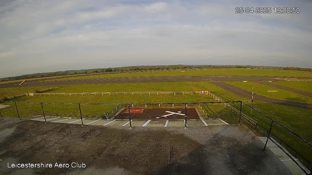 A wide view of an airfield taken from a webcam. In the foreground, there is a concrete platform with a railing, and a small staircase leading down to a paved area with marked parking spaces. Beyond the platform, there are green open fields stretching into the distance, with a runway running from left to right. The sky above is partly cloudy with patches of blue. The image also shows a small flag on a flagpole near the middle right and the date and time stamp at the top right corner reads "25-04-2025 19:00:50". The text "Leicestershire Aero Club" is visible in the bottom left corner.