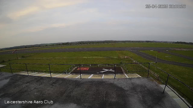 An empty outdoor airstrip or runway with grassy fields on both sides and a cloudy sky overhead. The image is taken from a high vantage point, possibly a building or control tower overlooking the airport. There are some markings and signs on the ground, and a few scattered objects along the runway. The timestamp in the upper right corner reads "25-04-2025 20:00:36," and the bottom left corner has the text "Leicestershire Aero Club."