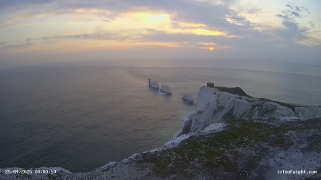 A view of white chalk cliffs along the coastline at sunset. The sky has soft clouds with hints of yellow, orange, and purple near the horizon. Ice formations are visible on the cliffs and in the sea below. A small structure is situated on top of the cliffs, with a path or railing running along the edge. The timestamp shows the date as April 25, 2025, at 8:00 PM.