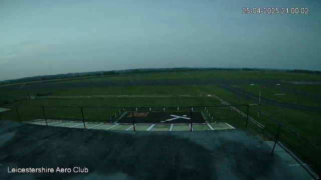 The image shows a view from a webcam overlooking a grassy airfield at LeicesterEshire Aero Club. The scene is outdoors under a cloudy sky, with a runway in the distance and a safety area with marked lines in the foreground. The timestamp in the top right corner indicates the date and time as April 25, 2025, at 21:00:02. The overall atmosphere is calm and overcast.