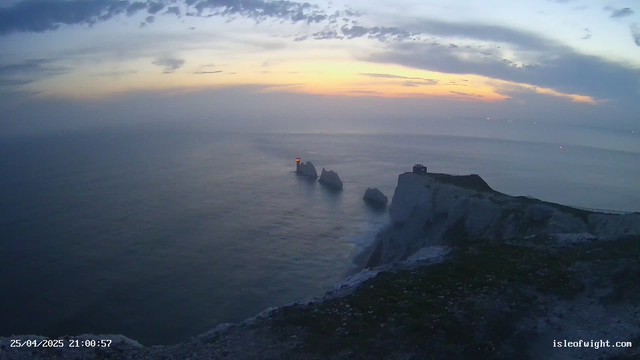 A view of the ocean with white chalk cliffs and several rock formations in the water. A lighthouse with a red light is visible on one of the rock formations. The sky is partly cloudy with the sun setting on the horizon, casting a soft light across the scene. The image includes a timestamp of "25/04/2025 21:00:57" and a website URL "isleofwight.com".