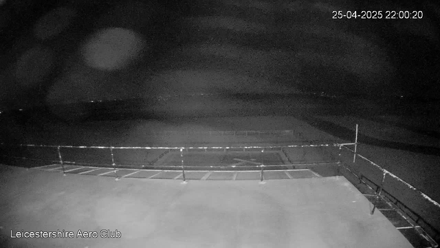 A black and white webcam image showing the exterior of Leicestershire Aero Club at night. The image features a tarmac area with a metal railing in the foreground, and open sky in the background with some distant lights faintly visible. The timestamp in the top right corner reads "25-04-2025 22:00:20."