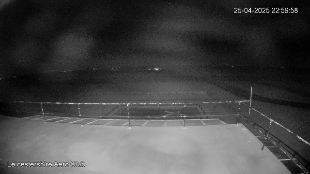 A black and white image from a webcam shows an empty outdoor area at night. There is a paved surface with a railing on the edge, possibly a viewing platform or open area. In the background, the sky is dark with a few faint stars, and distant lights are barely visible. The date and time are displayed in the top right corner as "25-04-2025 22:59:58." The bottom left corner indicates the location as "Leicestershire Aero Club."