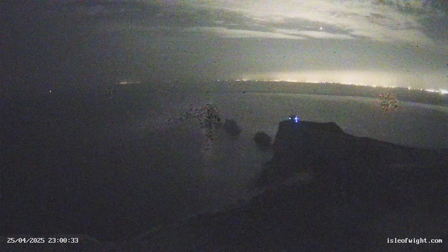 A dark, low-light image shows a landscape possibly taken at night. There is a faint glow on the horizon, suggesting city lights or lightning in the distance. In the foreground, there's a silhouette of a structure or vehicle with a small blue light. The scene appears to have a cloudy sky with some illuminated areas, and the image includes a timestamp of "25/04/2025 23:00:33" in the bottom left corner. The website "isleofwight.com" is in the bottom right corner.