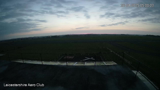 An outdoor scene taken from a webcam at Leicesterhire Aero Club shows an early morning sky with scattered clouds and a faint pink glow near the horizon. The landscape features expansive green fields stretching into the distance. In the foreground, part of a rooftop or platform with a safety railing is visible, along with some equipment or structures. The timestamp in the upper right corner indicates the date as April 26, 2025, and the time as 05:00:03.