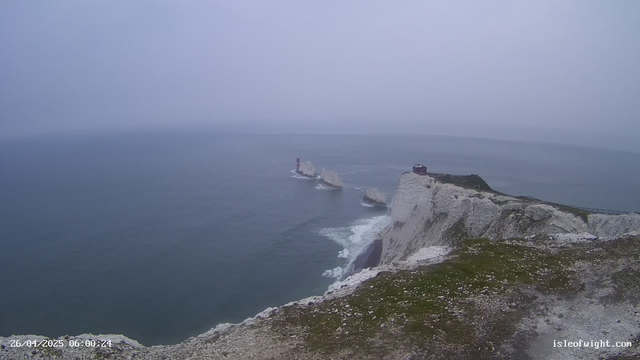 A foggy scene of white chalk cliffs overlooking the ocean, with a lighthouse perched on one of the cliffs. The sea is calm, and the sky is overcast, creating a moody atmosphere. A path runs along the top of the cliff, and some buildings are visible on the edge. The date and time stamp, "26/04/2025 06:00:24," is in the bottom left corner, and "isleofwight.com" is in the bottom right corner.