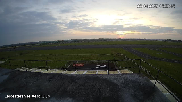 Early morning view of a large open field and runway at Leicestershire Aero Club, with a partly cloudy sky and the sun rising on the horizon. The time stamp shows 06:00:46 on April 26, 2025. There are railing and a helicopter landing pad in the foreground.