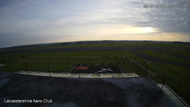 A view from a webcam at the Leicestershire Aero Club showing an early morning sky with partially cloudy clouds, a runway in the distance, and a helipad with a white helicopter symbol in the foreground. The timestamp reads 26-04-2025 07:00:32.