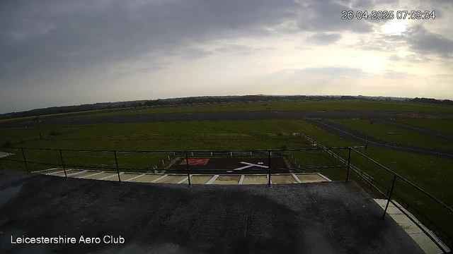 An outdoor view from a webcam at Leicestershire Aero Club. The foreground shows a black paved area with a railing, leading down to a grassy field with a red and white marker. Beyond the grass are runways and taxiways stretching into the distance under a cloudy sky, with the sun partially shining through. The timestamp in the upper right corner indicates the date and time as April 26, 2025, at 07:59:54.
