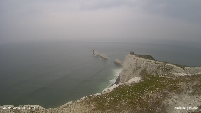 A view of white chalk cliffs with a grassy top extending into the ocean, under a gray, overcast sky. In the distance, a lighthouse or tower is situated among the rocks, and a small building is perched near the edge of the cliff. The water below is calm with gentle waves. The scene appears to be captured from a webcam, with a timestamp of "26/04/2025 08:01:02" at the bottom left corner.