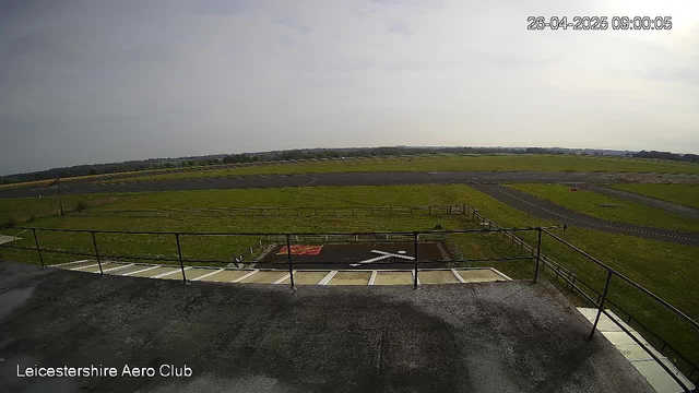 A wide view of an airfield shows a paved area with parking spaces in the foreground, a grassy field extending into the distance, and a runway to the far right. The sky is partly cloudy with sunlight casting shadows. The bottom left corner has the text "Leicestershire Aero Club."