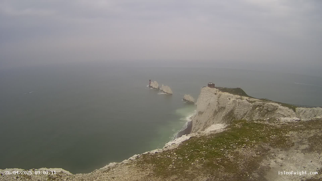 A view of white chalk cliffs extending into the sea with a small building or lighthouse on top. The ocean is calm with a few boats visible in the distance, and the sky is overcast with grey clouds.