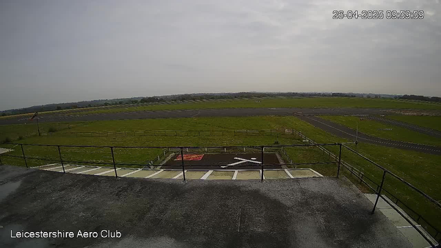 An outdoor view of a racing circuit with surrounding green fields and a cloudy sky. The image is taken from a high vantage point, showing part of the parking area with parking spaces and a railing in the foreground. The timestamp in the upper right corner reads "26-04-2025 09:59:59," and the bottom left corner has the text "Leicestershire Aero Club."