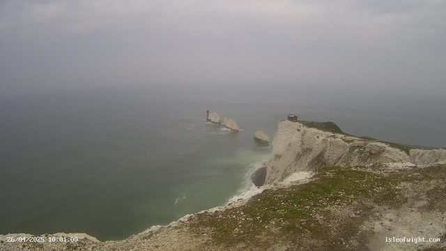 A panoramic view of white chalk cliffs extending into the ocean, with several large rocks near the shoreline and a lighthouse on one of the distant rocks. The sky is overcast, creating a gray and moody atmosphere. The cliffs are topped with some grassy areas and a walking path along the edge. The timestamp in the corner indicates the image was captured on April 26, 2025, at 10:01 AM.