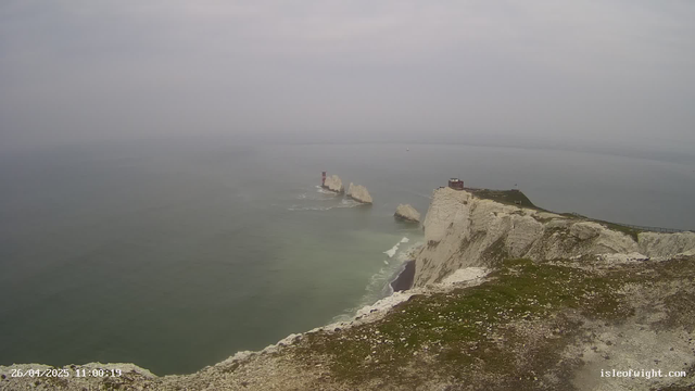 A view of white chalk cliffs extending into the ocean with a lighthouse situated on one of the cliffs. The coastline features steep cliffs with patches of grass on top, and large rocks are visible in the water near the cliffs. The sky is overcast, and the sea appears calm with gentle waves. In the bottom left corner, there's a timestamp, and in the bottom right corner, the website "isleofwight.com" is displayed.