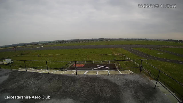 A view from a webcam at Leicestershire Aero Club showing an overcast sky, a grassy airfield with taxiways and runways, a small airplane in the distance, a white vehicle parked near the edge of the platform, and a small fenced area with a red marking on the ground. The timestamp in the top right corner reads "26-04-2025 11:00:29."