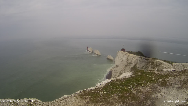 A view of white chalk cliffs along the coastline with a lighthouse and a small building on top. The sea below is calm with a few rocks protruding from the water. A boat speedily moves across the water, leaving a trail of white foam behind. The sky is overcast with gray clouds. The timestamp in the lower left corner indicates the date and time as 26/04/2025 at 12:00:53.