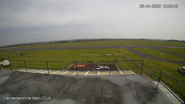 An outdoor view from a webcam showing an open grassy area with a racetrack or runway in the distance. In the foreground, there is a parking area with white lines and a fence, and a couple of vehicles parked on the right side. The sky is cloudy, and there is no visible activity or people. The timestamp in the upper right corner indicates the date and time as April 26, 2025, at 12:00:49. The bottom left corner has text that reads "Leicestershire Aero Club."