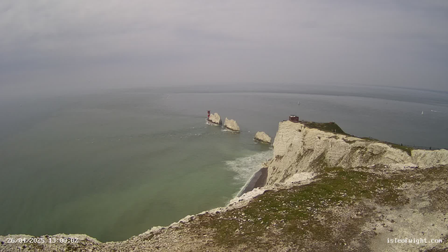 A view of high white chalk cliffs with a small building or observation point on top, extending into the ocean. Several large rock formations or stacks are visible in the water near the cliffs, with one having a red lighthouse or marker on top. The ocean appears calm, and there are sailboats scattered in the distance under a cloudy sky. The date and time are displayed in the lower left corner.