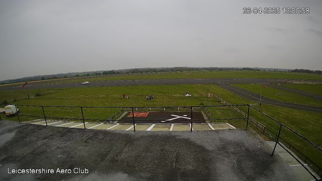 A wide view of an open grass field at Leicestershire Aero Club on a cloudy day. There is a small paved area with a railing in the foreground and some parked cars. In the distance, there are a few people near the grass. The landscape extends toward the horizon with a few roads visible. The timestamp in the upper right corner shows "26-04-2025 13:00:58."