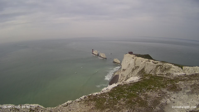 A panoramic view of white chalk cliffs rising from the sea, with a building on the top of the cliffs and small boats visible in the water below. The sky is overcast with gray clouds.