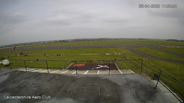 An outdoor view of Leicestershire Aero Club showing a grassy airfield with a paved runway stretching into the distance under a cloudy sky. There are a few small groups of people scattered on the grass near the runway, and a white vehicle parked on the left. In the foreground, there is a platform with a metal railing, and a designated area with painted markings on the ground. The date and time stamp in the top right corner reads "26-04-2025 14:00:58."
