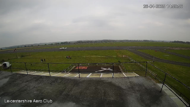 An overcast view of an airfield from a webcam, showing a grassy area with a few people sitting and standing, a small aircraft on the runway, and a parking area with marked spots in the foreground. The image includes the text "Leicestershire Aero Club" in the bottom left corner and a timestamp in the top right corner.