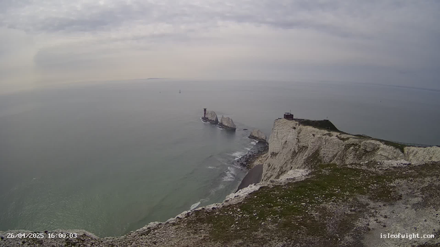 A view of the white chalk cliffs of Étretat on a cloudy day. The cliffs extend into the sea, with a small building and a lighthouse situated near the edge. The ocean appears calm with a few small sailboats visible in the distance. The foreground shows the rocky, grassy edge of the cliff. The timestamp at the bottom left indicates the date as April 26, 2025, at 16:00:03.