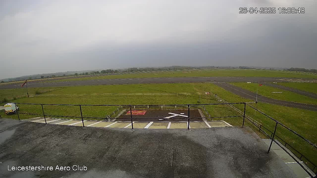 An empty open area at Leicestershire Aero Club with a parking lot in the foreground, a grassy field with a windsock and some fencing, and a runway extending into the distance under a cloudy sky. The timestamp on the image reads "26-04-2025 16:00:48."