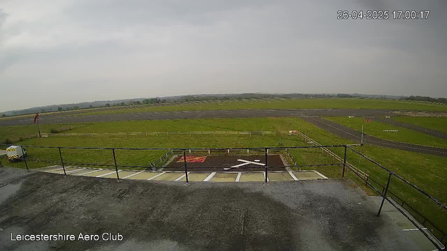 An outdoor view from a webcam showing a grassy area with a small paved section and a white arrow painted on the ground, pointing to the right. in the background, there are open fields with roads, and the sky is cloudy. The image is labeled "Leicestershire Aero Club," and the date and time are displayed in the top right corner as "26-04-2025 17:00:17."