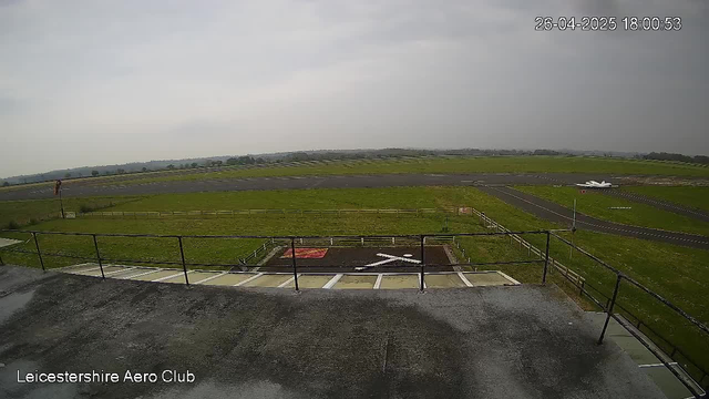 An overcast sky over a small airfield with a short runway. In the foreground, there is a balcony with a black railing and parking spaces below it. A white aircraft is taxiing on the runway in the distance. The image has a timestamp in the upper right corner and the label "Leicestershire Aero Club" in the lower left corner.