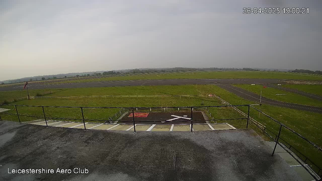 An empty airfield with a parking area in the foreground, a grassy runway, and open fields in the background under a cloudy sky. The timestamp in the top right corner reads "26-04-2025 19:00:21," and "Leicestershire Aero Club" is written in the bottom left corner.
