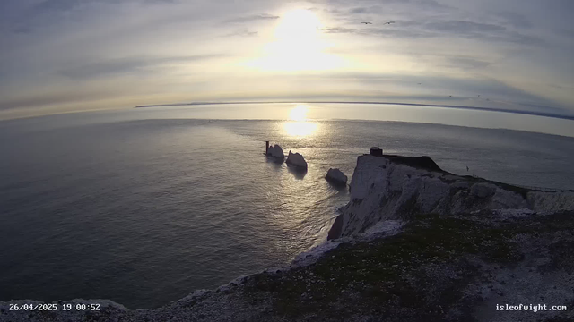 A view of a coastal landscape during sunset or sunrise, featuring the white cliffs of Étretat with a rock formation in the water. The sky has some thin, wispy clouds, and the sunlight reflects off the calm sea. The timestamp in the corner indicates the date and time as April 26, 2025, at 19:00:52.