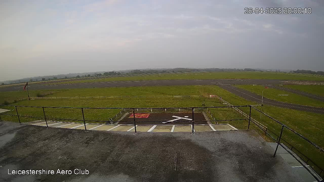 A view of an airfield with a parking area in the foreground, including a helicopter landing pad marked with an "H" and white lines. Beyond the parking area, there is a grassy expanse with runway markings, taxiways, and a distant horizon with rolling hills. The sky is mostly cloudy with some patches of blue. The image is timestamped "26-04-2025 20:00:10" in the top right corner, and the bottom left corner has a label reading "Leicestershire Aero Club."
