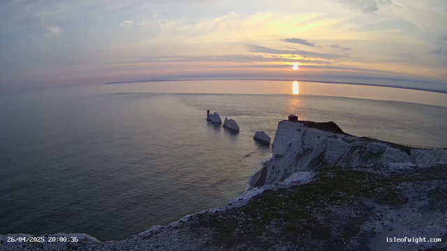 A stunning sunset over the ocean with the sky filled with soft clouds. White chalk cliffs are visible in the foreground, with a small structure and a railing at the edge. Several large white rocks or sea stacks emerge from the water near the cliffs. The reflection of the setting sun gleams on the calm water. The image is marked with a timestamp of April 26, 2025, at 8:00 PM, and the website "isleofwight.com" is displayed at the bottom right corner.