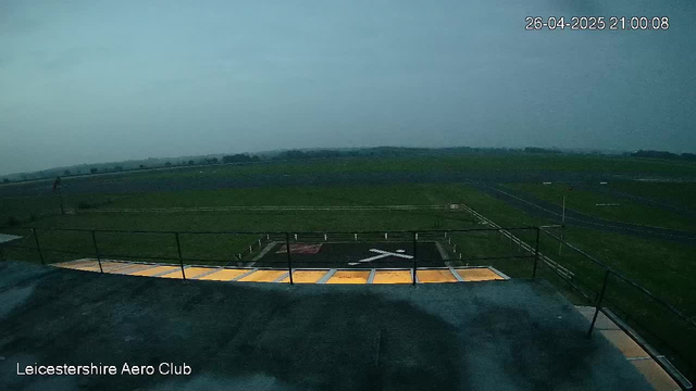 An empty helicopter landing pad with yellow markings and a white "X" in the center, surrounded by a safety railing, is viewed from a vantage point at the Leicesterhire Aero Club. The landscape extends into open fields under a cloudy sky, and the timestamp in the top right corner reads "26-04-2025 21:00:08."