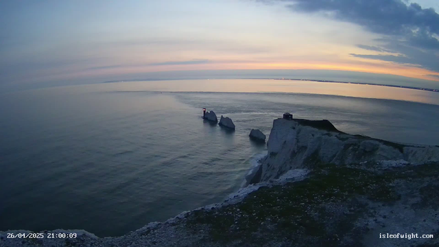 A view of a coastal landscape at dusk, featuring white chalk cliffs with a small building on top, a group of rocks in the water, and a lighthouse on one of the rocks. The sky displays soft pastel colors with the sun setting on the horizon. The calm sea reflects the sky's colors. A timestamp and website are visible in the bottom corners.