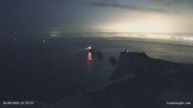 A nighttime scene of a coastal area with a calm body of water. In the distance, there are faint lights along the shoreline and a bright glow illuminating the horizon. There are rocks or small structures partially submerged in the water in the foreground, with some small lights visible on or near them. The overall atmosphere is dark, with minimal artificial lighting, and the sky appears overcast or cloudy. The timestamp in the bottom left reads "26/04/2025 21:59:51," and in the bottom right, there is a website URL: isleofwight.com.