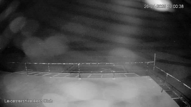 A black-and-white webcam image shows an empty outdoor area at night, with a railing or barrier edge at the bottom. The sky appears cloudy, and no people or vehicles are visible. The timestamp in the upper right corner reads "26-04-2025 22:00:38." The words "Leicestershire Aero Club" are displayed in the lower left corner.